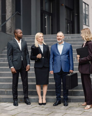 Confident businesspersons standing in front of modern office building. Men and women talking and holding documents. Banking and financial market concept.