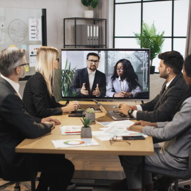 Confident Caucasian business man and African woman tells business program concept to their multinational colleagues during a video conference in a modern office with a huge TV for conversations